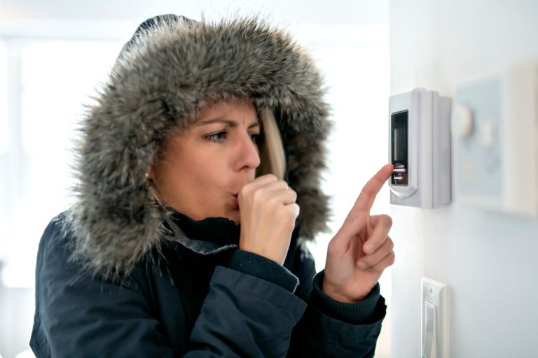 woman wearing a winter coat indoors adjusting her broken heating thermostat