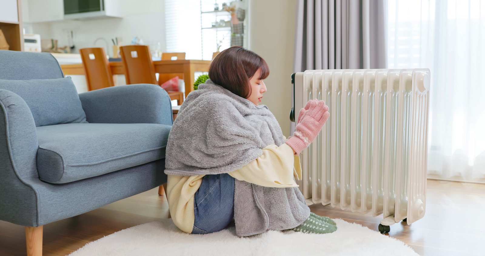 A girl wrapped in a thick grey blanket sitting on the floor next to a radiator to stay warm.