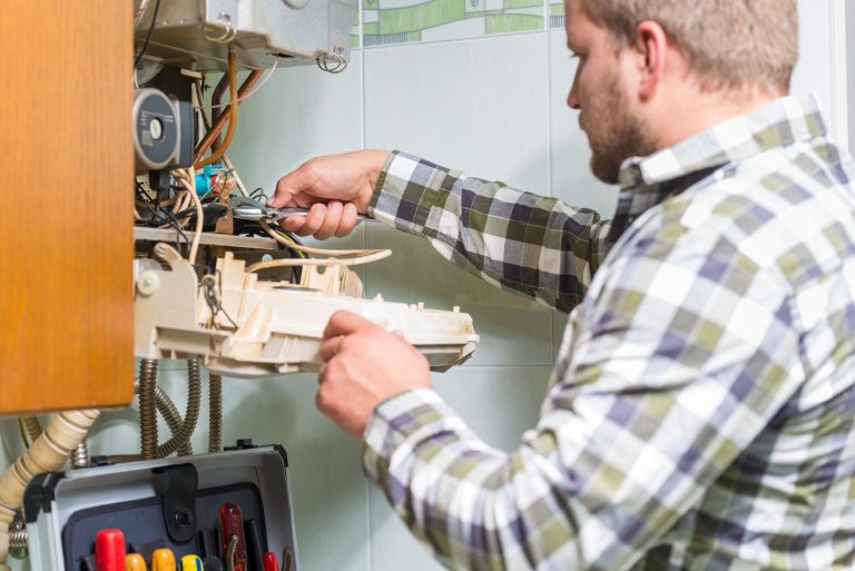 A technician in a plaid shirt working on the internal components of a wall-mounted boiler.