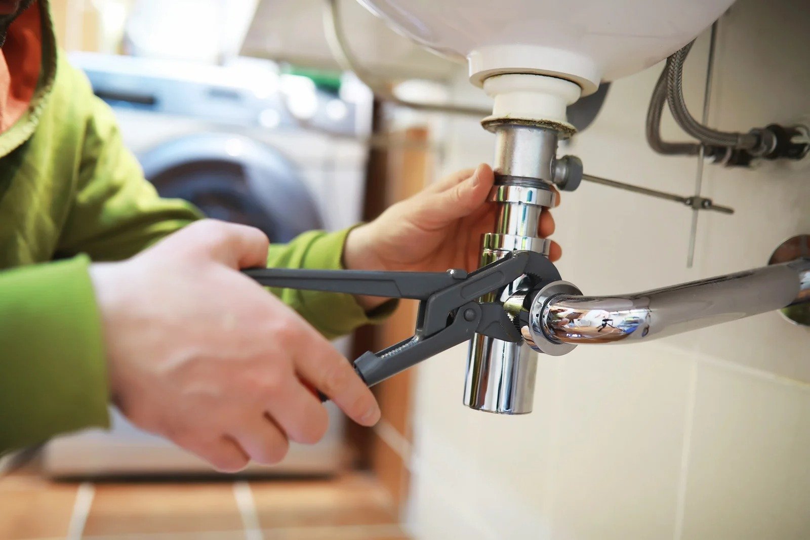 a man in a green sweater using a wrench on a water pipe below a bathroom sink