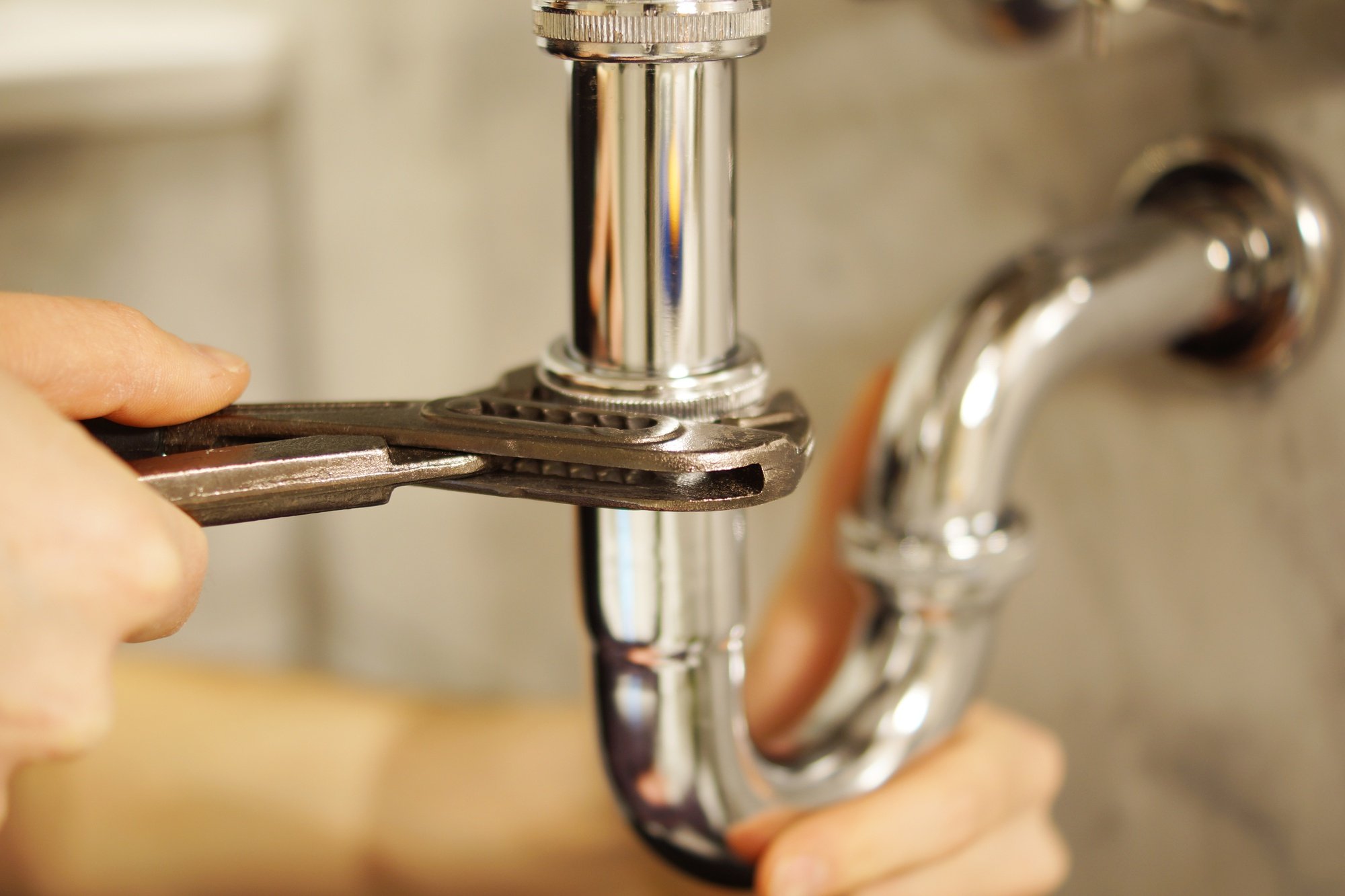 Close-up of a chrome P-trap pipe under a kitchen sink being tightened with a silver adjustable wrench.