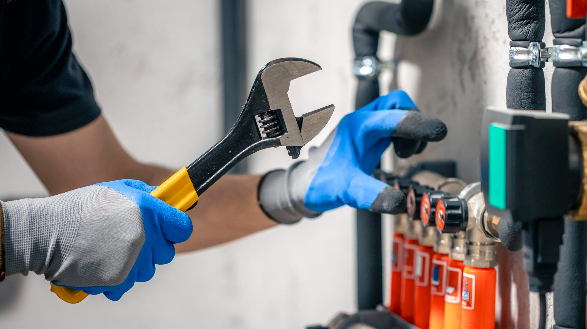 A plumber wearing blue protective gloves uses a large wrench to work on a complex manifold system with red and blue temperature gauges.
