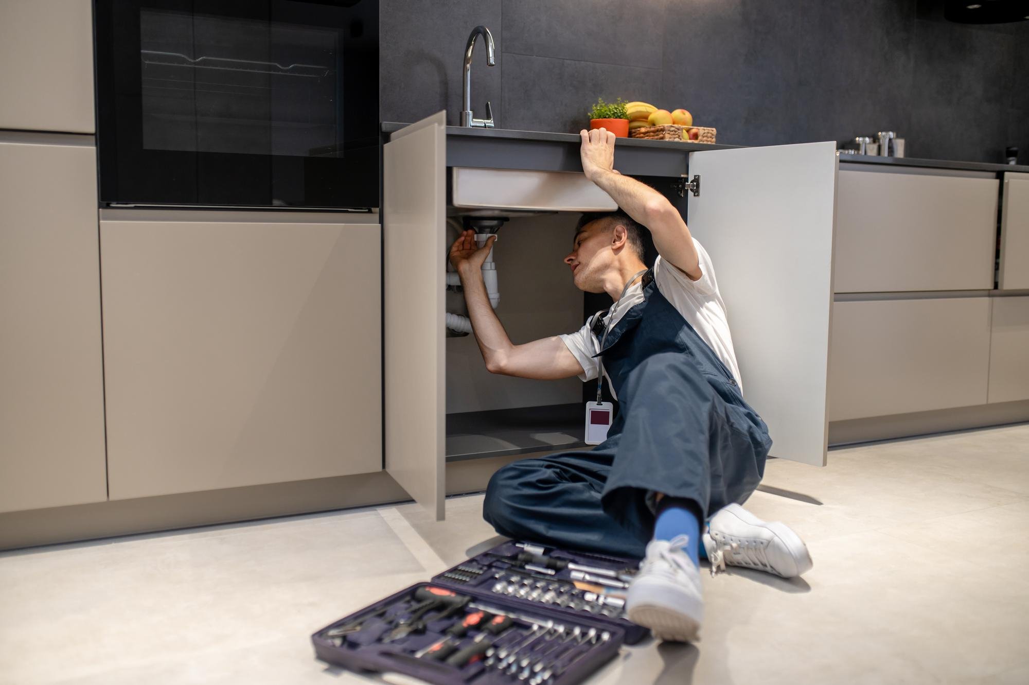 A plumber in overalls works under a kitchen sink with open cabinet doors, surrounded by a toolbox
