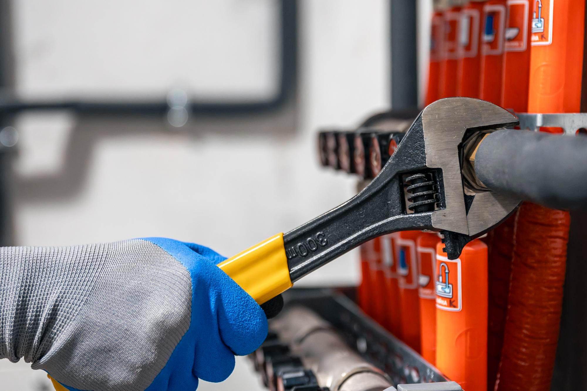 Close-up of a gloved hand using a wrench on a black pipe connected to a bright orange vertical tank or radiator system.