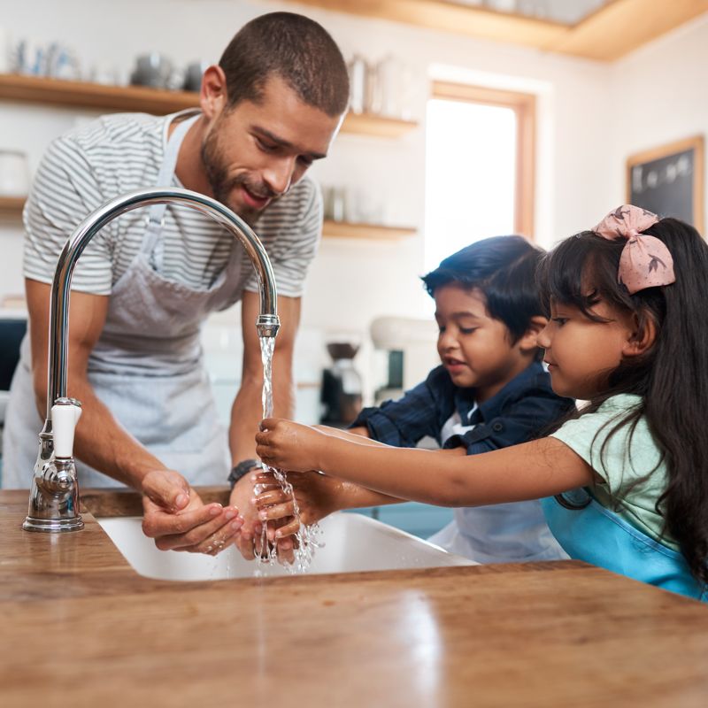 A man wearing an apron helps two young children wash their hands under a kitchen faucet.