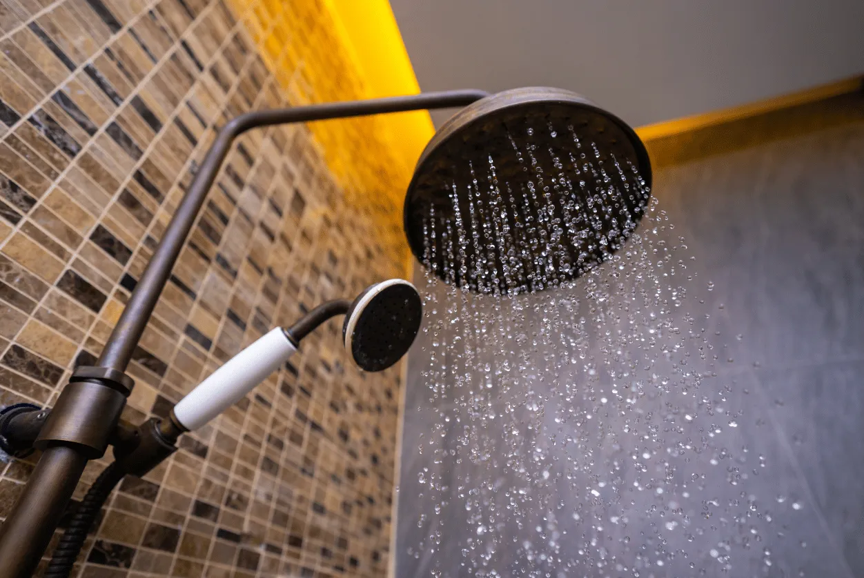 Close-up of a dark metal shower head with water flowing, next to a handheld shower. The background features tiled walls and warm lighting.